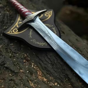 Könnte beinhalten: Ein detailliertes Schwert mit einer silbernen Klinge und einem rot-weiß gemusterten Griff. Das Schwert liegt auf einer Holzplakette mit goldenen Akzenten und Inschriften. Die Klinge hat Inschriften entlang ihrer Kante. Das Schwert wird auf einer strukturierten Holzoberfläche präsentiert.