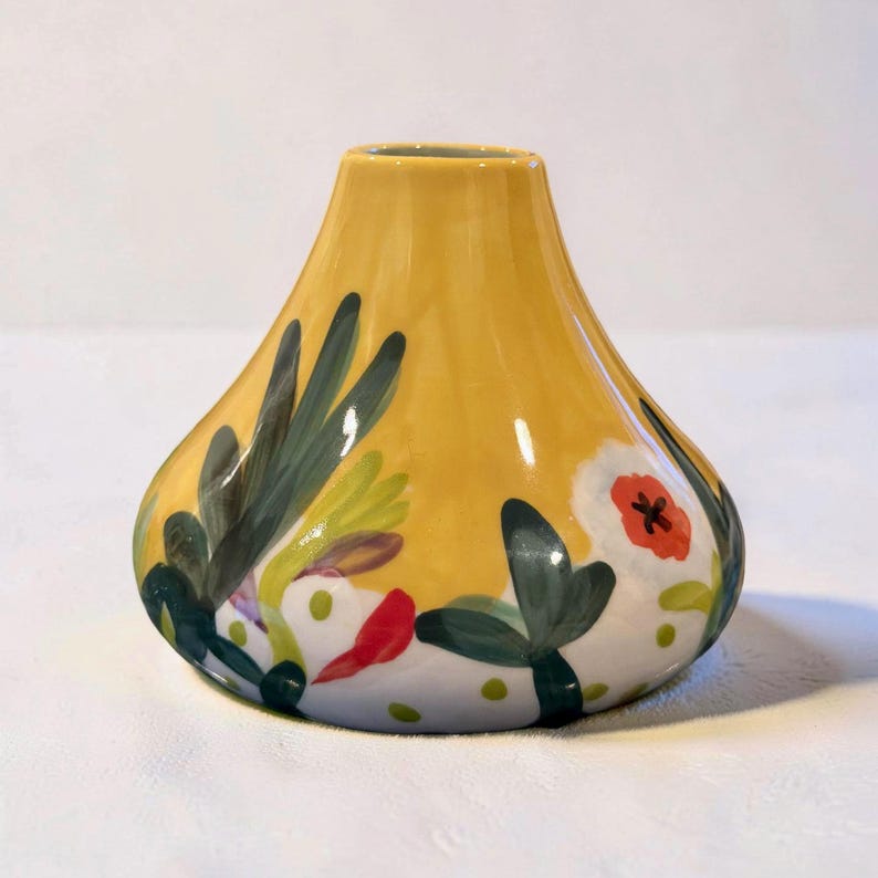 May include: A yellow ceramic vase with a bulbous base and a narrow neck. The vase is decorated with hand-painted floral and foliage designs in green, red, and white. The vase is approximately 7 inches tall.