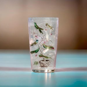 May include: A clear glass vase with a pink tint and a design of green and blue birds on a branch with pink flowers and a nest at the bottom.