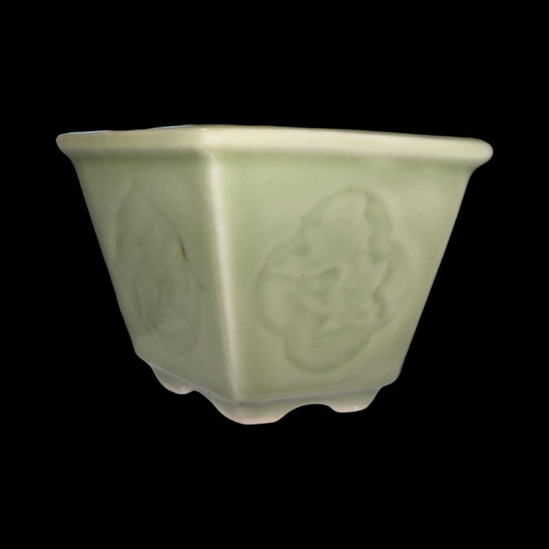 May include: A light green ceramic planter with a square shape and a scalloped rim. The planter has a raised design of stylized flowers on each side.