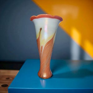 May include: A tall, elegant glass vase with a flared, ruffled rim. The vase features a white body with orange and yellow leaf-like patterns. The base and rim are accented with a metallic orange hue, creating a striking contrast. The vase is a decorative home accent.