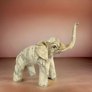 May include: A white ceramic elephant sculpture with a long trunk raised in the air. The elephant has a textured surface and a detailed face.