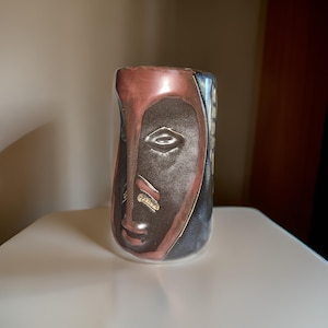 May include: A ceramic vase with a stylized face design. The vase is brown, black, and red with gold accents.