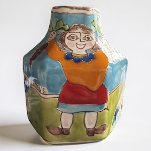 May include: A hexagonal ceramic vase featuring a hand-painted illustration of a figure with brown hair, wearing an orange top, red skirt, and blue skirt over light-colored legs with brown shoes, set against a blue sky and green grass background.