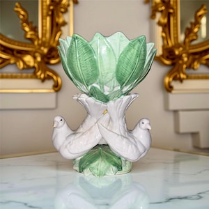 May include: A decorative ceramic centerpiece featuring a green leaf-shaped bowl atop a white base with two doves. The doves are perched on a base of green leaves. The background includes gold-framed mirrors.