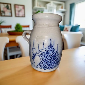 May include: A stoneware pitcher with a handle, featuring a blue and white design of a figure decorating a Christmas tree. The pitcher has a wide mouth and a slightly textured surface. The design is hand-painted, giving it a rustic, vintage feel.