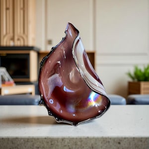 May include: A decorative glass sculpture in shades of plum, white, and iridescent hues. The freeform shape has a ruffled edge with a silver trim. The glass reflects light, creating a dynamic visual effect. This art piece is suitable for home decor.