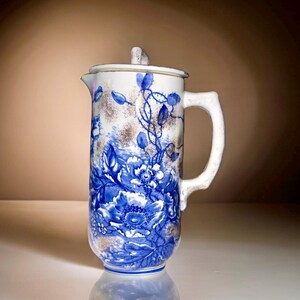May include: A white ceramic pitcher with a blue and white floral pattern. The pitcher has a lid and a handle.