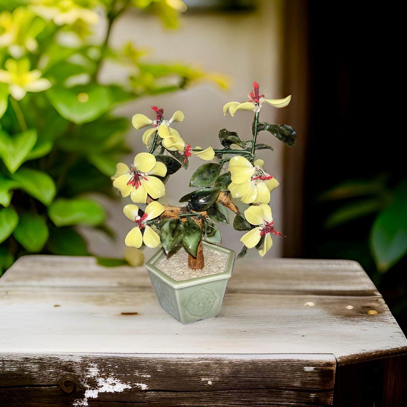 May include: A small glass bonsai tree with yellow flowers and green leaves in a green ceramic pot. The bonsai is on a wooden table.