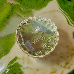May include: A small, green ceramic dish with a scalloped edge and a swirling pattern. The dish is glazed in a light green color with a hint of brown.