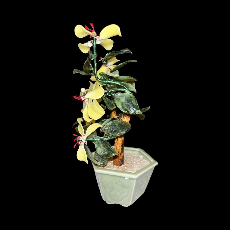 May include: A jade bonsai tree with yellow flowers in a green ceramic pot. The tree is made of green jade and has a brown wooden trunk. The flowers are made of glass and are yellow with red centers.