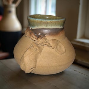 May include: Handmade ceramic vase with a bulbous body and a wide opening. The vase features a textured, light brown surface with sculpted relief details. The rim is glazed with shades of green and brown. A second vase is blurred in the background.