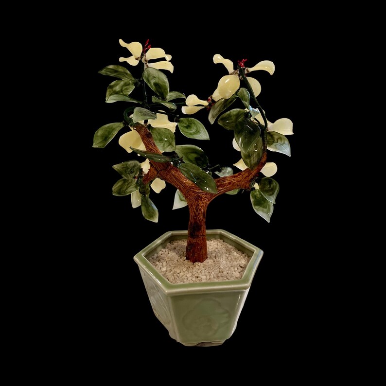 May include: A jade bonsai tree with white flowers and green leaves in a green ceramic pot. The tree is made of jade and the pot is decorated with a floral design.