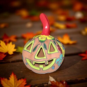 May include: A ceramic jack-o'-lantern with a brown base, green and yellow accents, and a red stem. The pumpkin has triangular cutouts for eyes and a carved smile. Decorative floral patterns are painted around the pumpkin's face. The background features fall leaves.