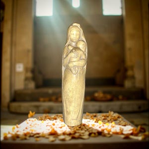 May include: A golden-toned statue of a mother holding a child. The sculpture is set against a blurred background with sunlight streaming through windows. The statue is on a stone surface with scattered leaves.