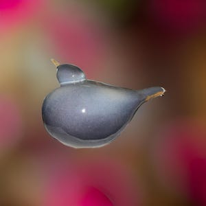 May include: A small, gray ceramic bird figurine with a rounded body and a long, thin beak. The bird is perched on a blurred background of pink and orange.