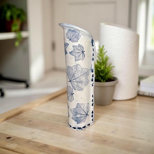 May include: A tall, off-white ceramic vase with a curved design. The vase features a pattern of blue leaf imprints. The edge is trimmed with blue dots. The vase is approximately 12 inches tall.