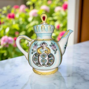 May include: A decorative ceramic teapot with a detailed hand-painted design. The teapot features a light blue and yellow color scheme with floral and figure motifs. The handle and spout are white, and the lid has a red accent.