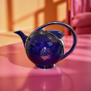 May include: A navy blue ceramic teapot with a gold handle and trim. The teapot features gold floral designs and a curved handle. The teapot is sitting on a pink surface.