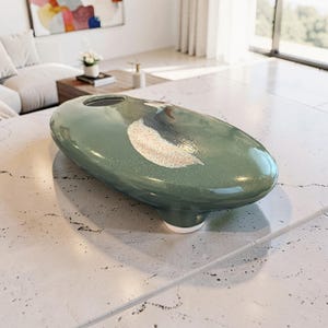 May include: A green ceramic vase with an oval shape and a single opening. The vase has a white and gray abstract design on the side. The vase is a decorative item for a home.