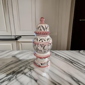 May include: A white ceramic jar with a pink and blue floral design. The jar has a lid and a base with a scalloped edge. The jar is sitting on a marble surface.