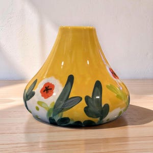 May include: A yellow ceramic vase with a wide base and a narrow neck. The vase is decorated with hand-painted floral designs, including white flowers with red centers and green leaves. The vase is sitting on a wooden surface.