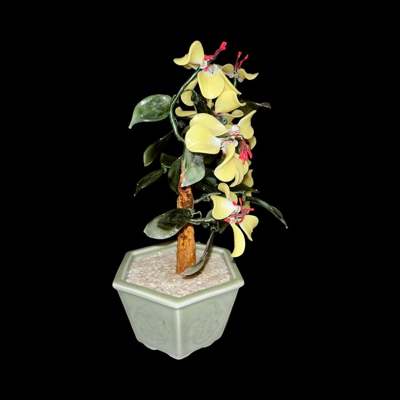 May include: A jade bonsai tree with yellow flowers and a green ceramic pot. The tree is made of jade and has a brown wooden trunk. The pot is hexagonal and has a white glaze.