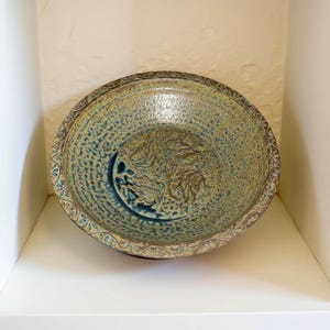 May include: A ceramic bowl with a textured, speckled surface in shades of blue and green. The bowl features a detailed leaf design in the center and a decorative border. The bowl is displayed on a white shelf.