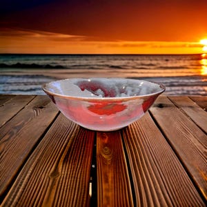 May include: A large, white ceramic bowl with a red glaze. The bowl is sitting on a wooden surface. The background is a blurry image of a sunset over the ocean.