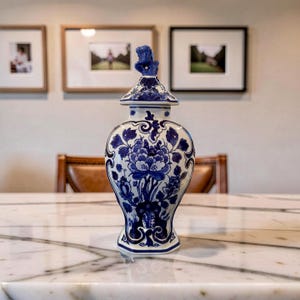 May include: A white and blue ceramic decorative jar with a lid. The jar features a floral design in blue and white. The lid has a blue floral finial. The jar is sitting on a marble surface.