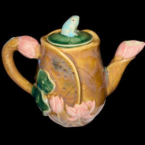 May include: A brown ceramic teapot with a frog on the lid and lotus flower designs. The teapot has a green leaf design on the lid and a pink lotus flower design on the spout.