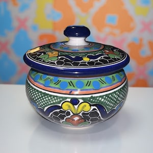 May include: A hand-painted ceramic jar with a lid. The jar is round with a blue rim and lid, and is decorated with green, blue, orange, and yellow floral patterns. The jar has a white base and a dark blue knob on the lid.