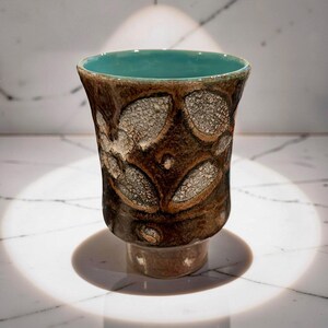 May include: A handmade ceramic cup with a textured, brown exterior and a turquoise interior. The cup features a unique pattern of raised, oval-shaped designs. The base is a lighter brown, and the cup is approximately 4 inches tall.