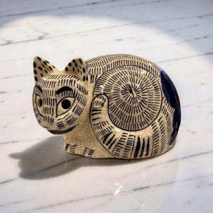 May include: A ceramic cat figurine in a curled-up position. The cat is cream-colored with intricate black line detailing and dark blue accents. The eyes are black, and the ears have black spots. The figurine has a crackle glaze finish.