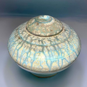 May include: A ceramic lidded jar with a light blue and white glaze. The jar has a round body and a wide, flat lid. The glaze has a mottled effect, with streaks of blue and white.