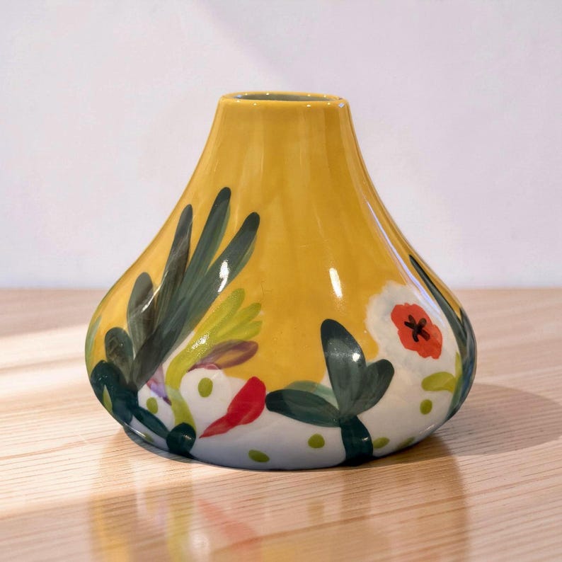 May include: A yellow ceramic vase with a bulbous shape and a narrow neck. The vase is decorated with hand-painted floral and foliage designs in green, red, and white. The vase sits on a light-colored wooden surface.