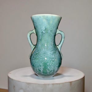 May include: A teal-colored ceramic vase with two handles. The vase has a bulbous base and a flared top. The surface has a speckled, textured appearance. The vase is displayed on a white, cylindrical pedestal.