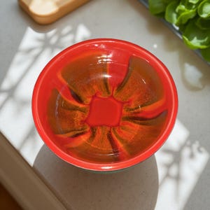 May include: A vibrant, round ceramic bowl with a glossy finish. The bowl features a bright red exterior and a striking interior design of orange and brown hues. The bowl's design is centered with a square shape. The bowl is suitable for serving food.