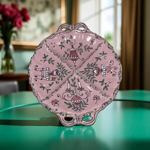 May include: A pink ceramic serving dish with a scalloped edge and four sections. The dish is decorated with green leaves, red and white lanterns, and other floral motifs. The dish has two handles.