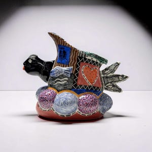 May include: A whimsical ceramic sculpture in the shape of a fish. The black fish has a red tongue and is decorated with a patchwork of patterns, including a heart, fish, and a blue figure. The base is a mix of pink, blue, and purple spheres.