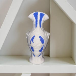 May include: White ceramic vase with blue accents. The vase has a unique shape with a flared top and decorative blue patterns. The vase is sitting on a white shelf.