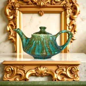 May include: A ceramic teapot with a turquoise and brown glaze. The teapot has a curved spout, a rounded body, and a handle. The lid is green. The teapot is displayed on a white surface with a gold ornate frame in the background.