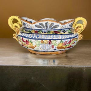 May include: A white ceramic bowl with yellow handles and colorful floral and geometric designs. The bowl is decorated with blue, red, green, and gold accents.