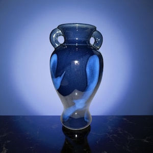 May include: A dark blue ceramic vase with two handles and a glossy finish. The vase features lighter blue, flowing patterns. The vase is on a dark surface with a blue gradient background.