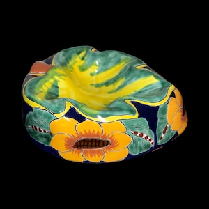 May include: A ceramic ashtray with a yellow, green, and blue floral design. The ashtray is shaped like a leaf and has a deep well in the center.