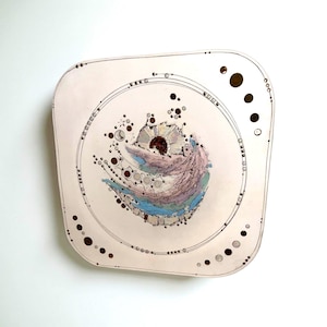 May include: A square, off-white ceramic plate with a central abstract design. The design features a swirling pattern of blue, purple, and brown hues, surrounded by a circular line and dotted accents. The plate has rounded corners and a minimalist aesthetic.