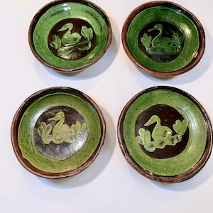 May include: Four ceramic bowls with a green and brown color scheme. Each bowl features a duck design in the center, surrounded by green and brown concentric circles. The bowls have a rustic, handmade appearance.