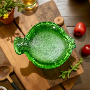 May include: A vibrant green glass dish with a textured surface, shaped like a leaf. The dish is set on a wooden cutting board, with a few red tomatoes and sprigs of greenery nearby. The dish is ideal for serving appetizers or small dishes.