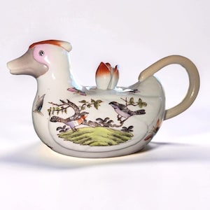 May include: A white ceramic teapot shaped like a duck, featuring a bird and floral design. The teapot has a curved handle and a spout resembling a duck's head. The lid is a flower bud. The teapot is decorated with colorful birds and butterflies.