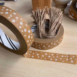 May include: Two rolls of brown paper tape with white polka dots. The tape is rolled up and the ends are visible. The tape is on a wooden surface.